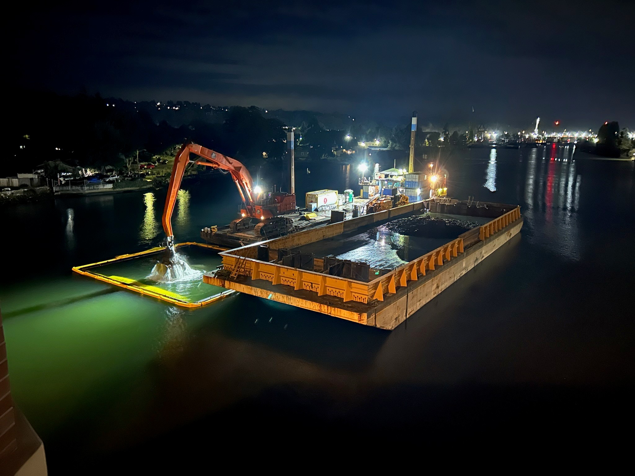Photo looking north on the waterway taken at night. Photo shows barge and dredge equipment. Dredge is pulling sediment out of the water while illuminated by light.
