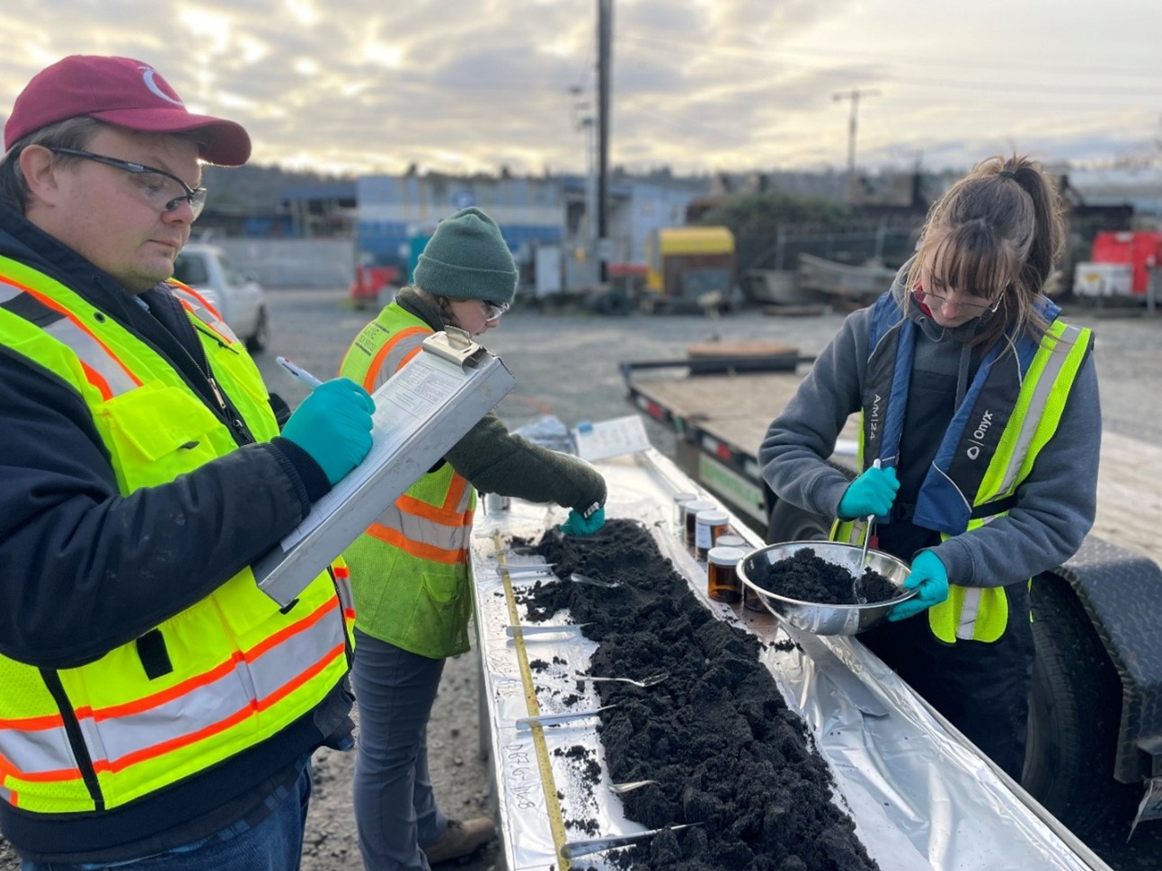 Photo of three people wearing safety vests. The people are looking at sediment core samples on a table. They are outside.