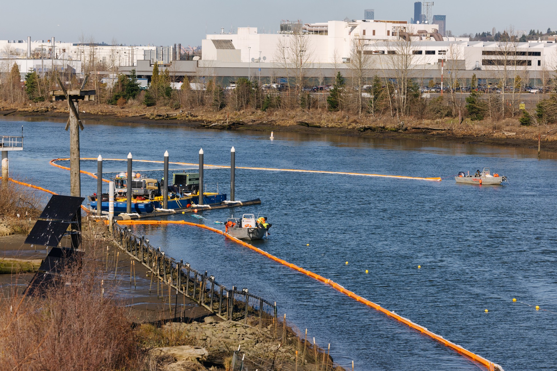 Photo showing sheen recovery crews adjusting and maintaining oil-containment booms and bubble curtain used to capture and manage sheens observed as a result of dredging SMA 12B. Image shows three vessels, a boom on the water, and building in background. Looking north.